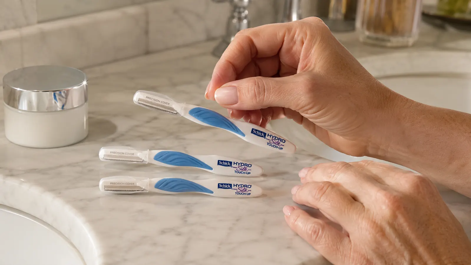 Woman's hand holding a small stack of three Schick Hydro Silk dermaplaning tools next to a makeup bag on a bathroom counter