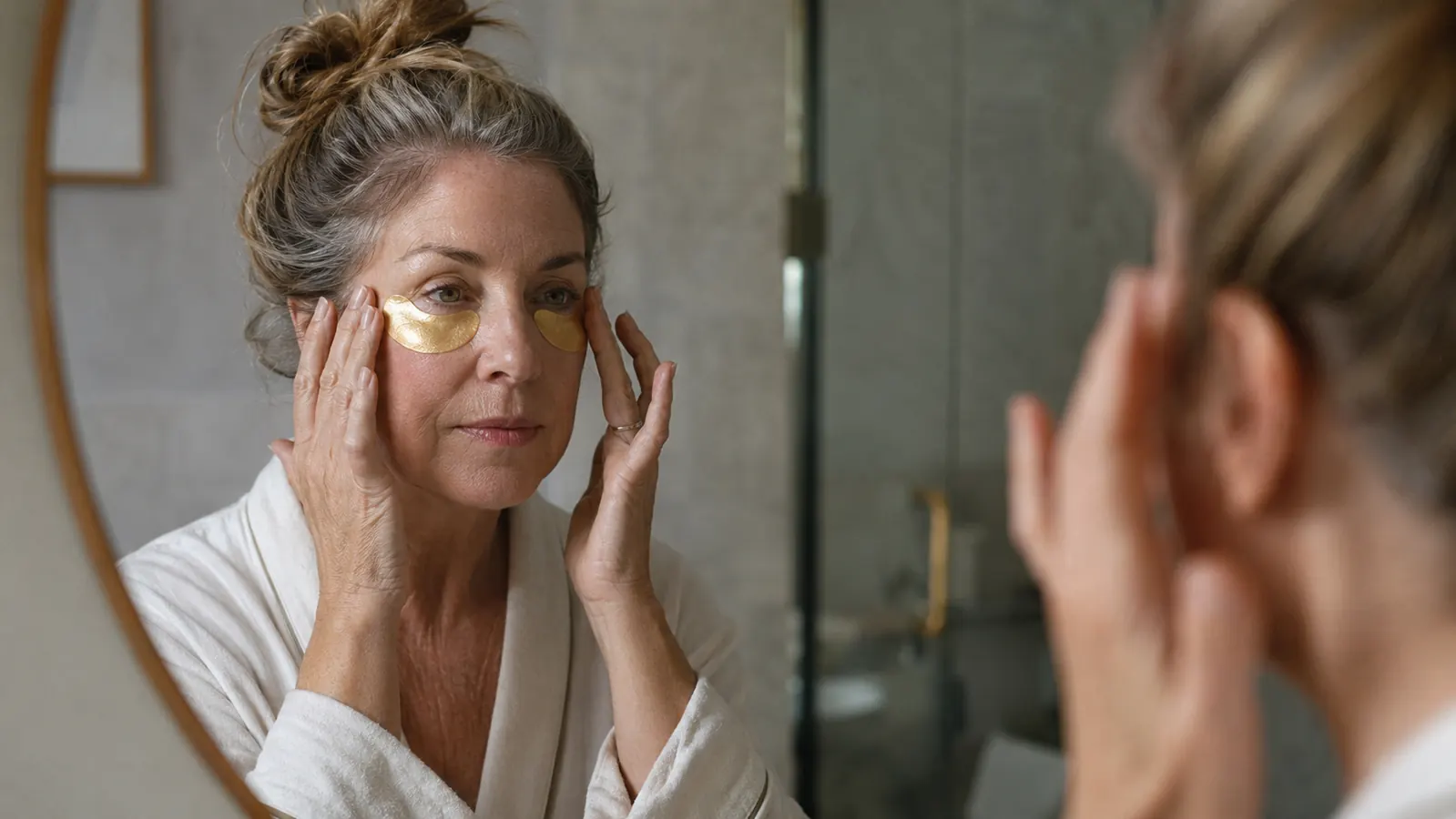 Woman at her bathroom mirror in soft morning light, pressing gold hydrogel patches under her eyes
