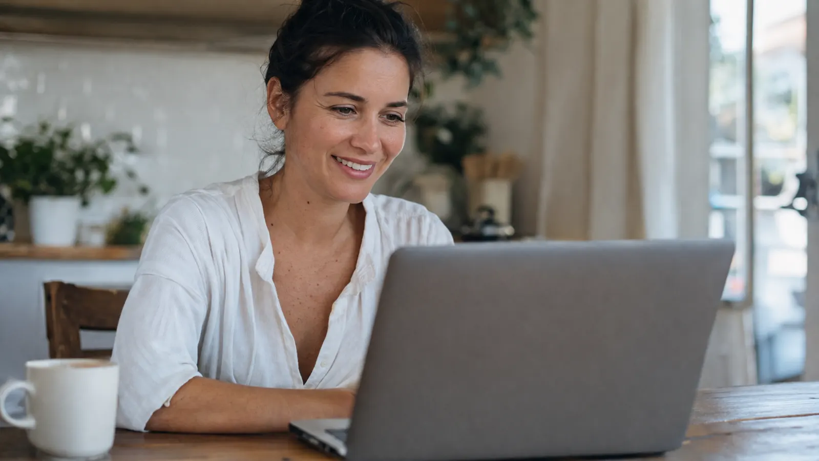 Woman smiling and refreshed at her kitchen table, ready for a video call with coffee in hand