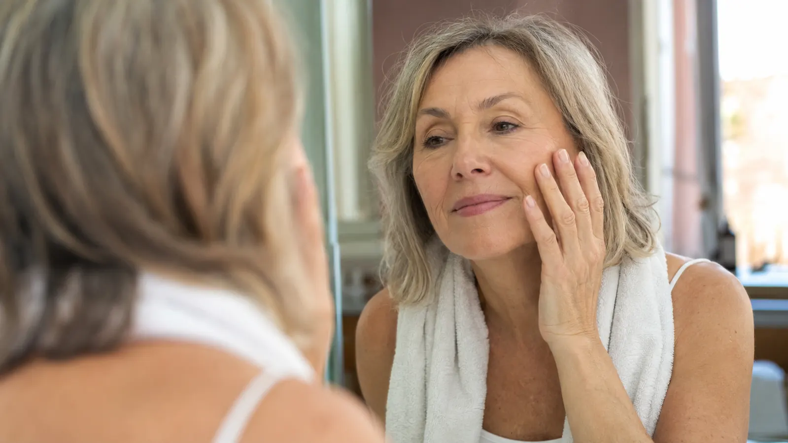 Older woman standing at a bathroom mirror in soft morning light, gently touching her smooth cheek