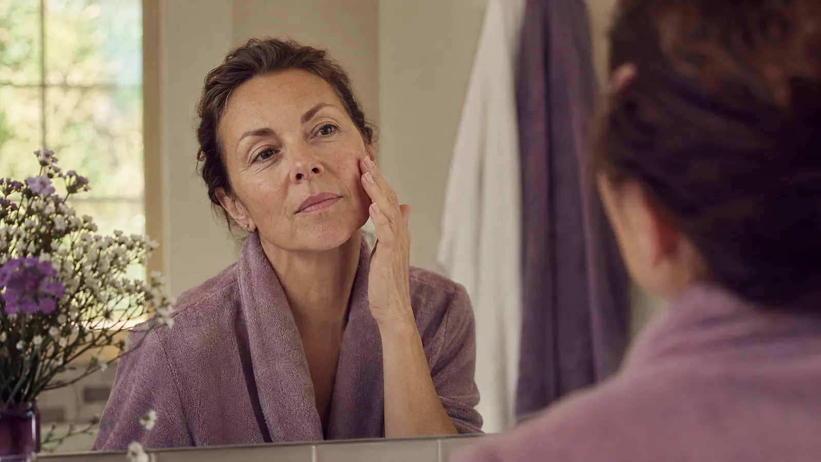 Close-up of a woman in her 50s gently pressing facial oil into her cheek, soft morning light, smooth glowing skin
