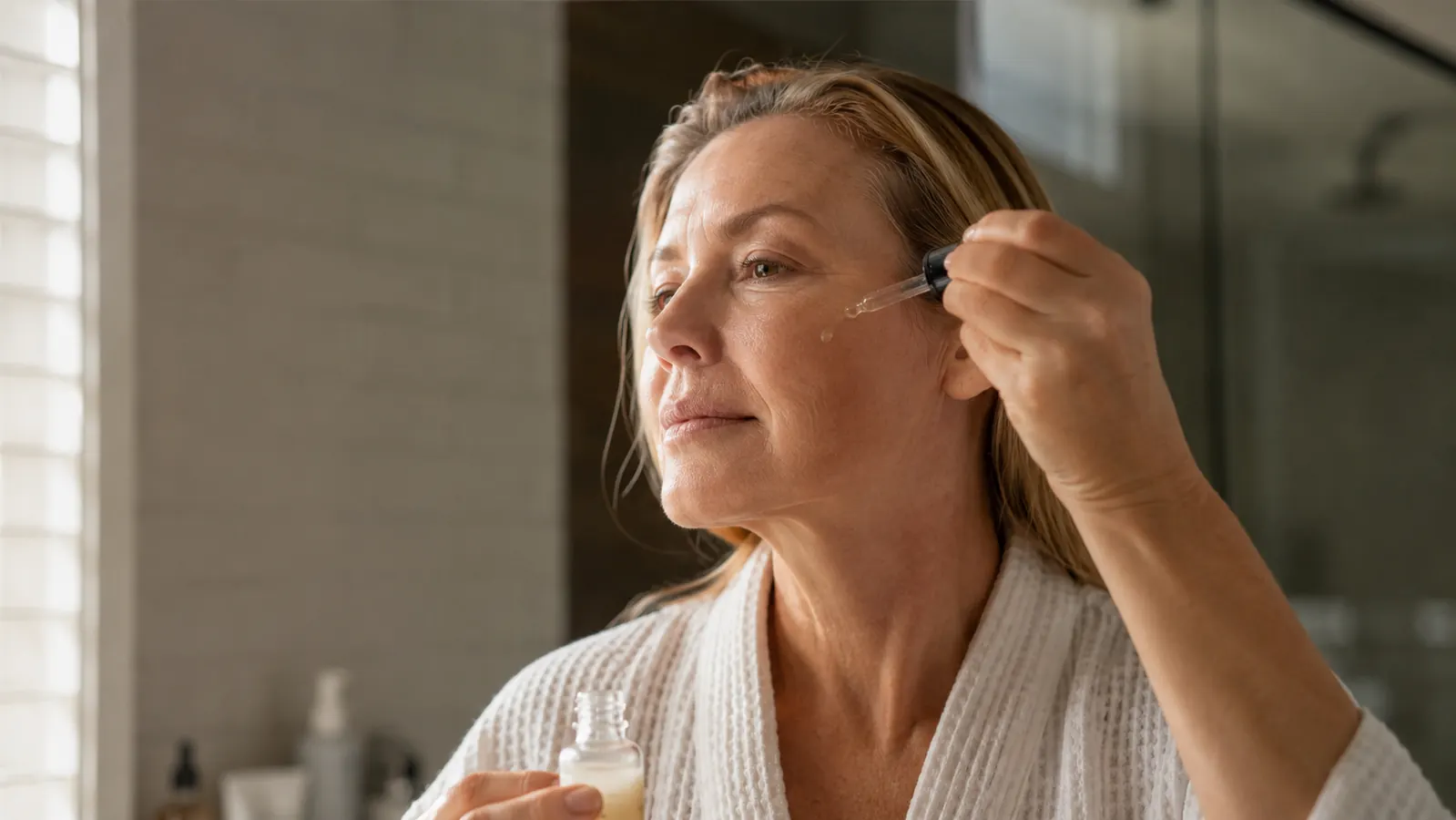 Woman in her 50s applying serum to her cheek in soft morning light at a bathroom mirror