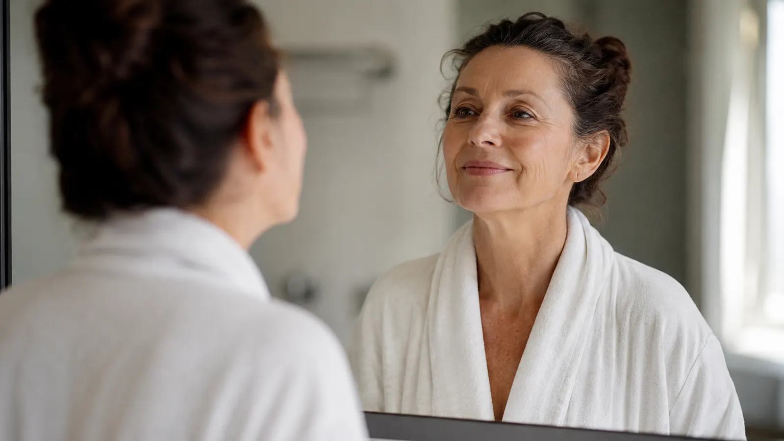 Woman in her 60s looking at her reflection in a bathroom mirror, soft morning light, calm and unposed