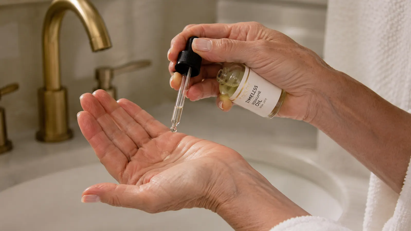 A hand pressing two drops of squalane oil into the palm before applying to the face, dropper bottle in the other hand