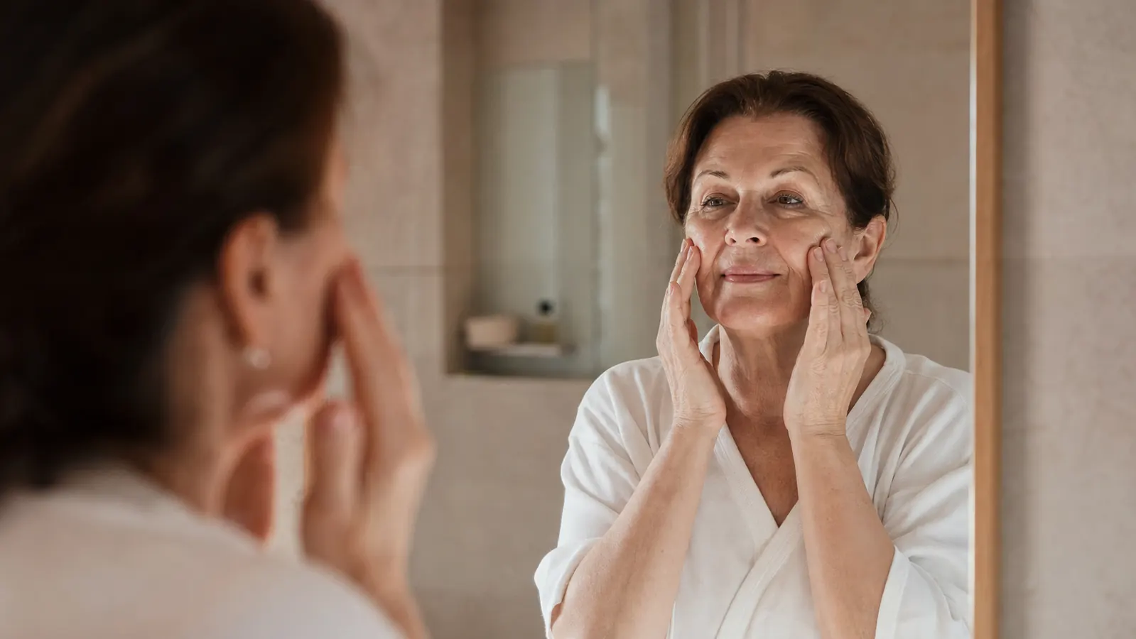 Woman in her 60s applying facial oil to glowing skin in soft morning light